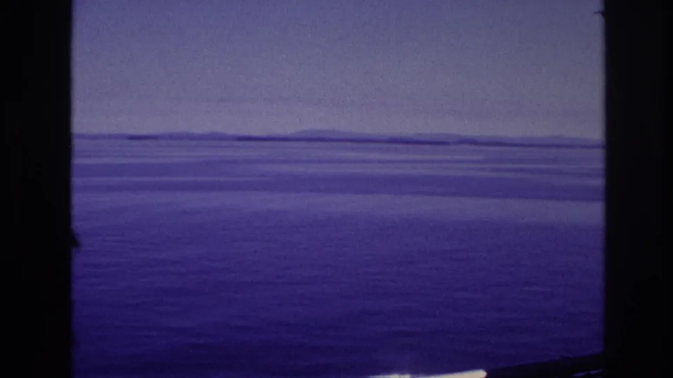 Shipboard 8mm view of calm water and a low distant shoreline under a hazy sky in the supplied 1967 Canada context.
