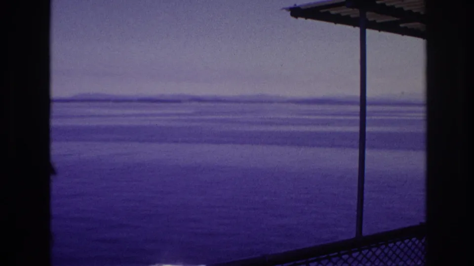 1967 8mm shipboard frame showing blue water, distant land, a deck overhang, railing, and vertical support.