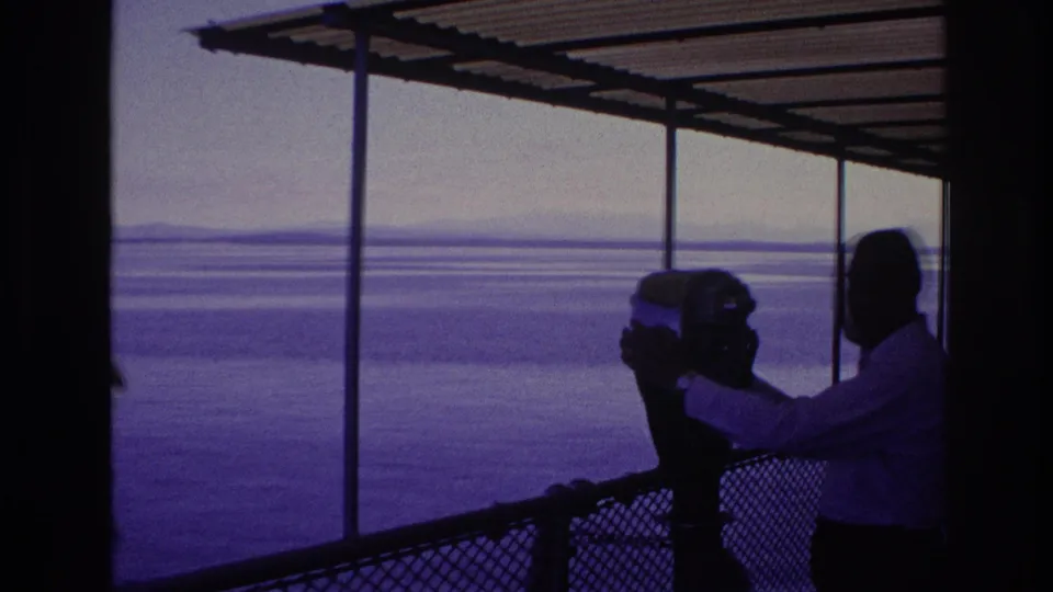A silhouetted adult man in a light shirt stands at a coin-operated telescope on a covered waterfront overlook in 1967 Canada.