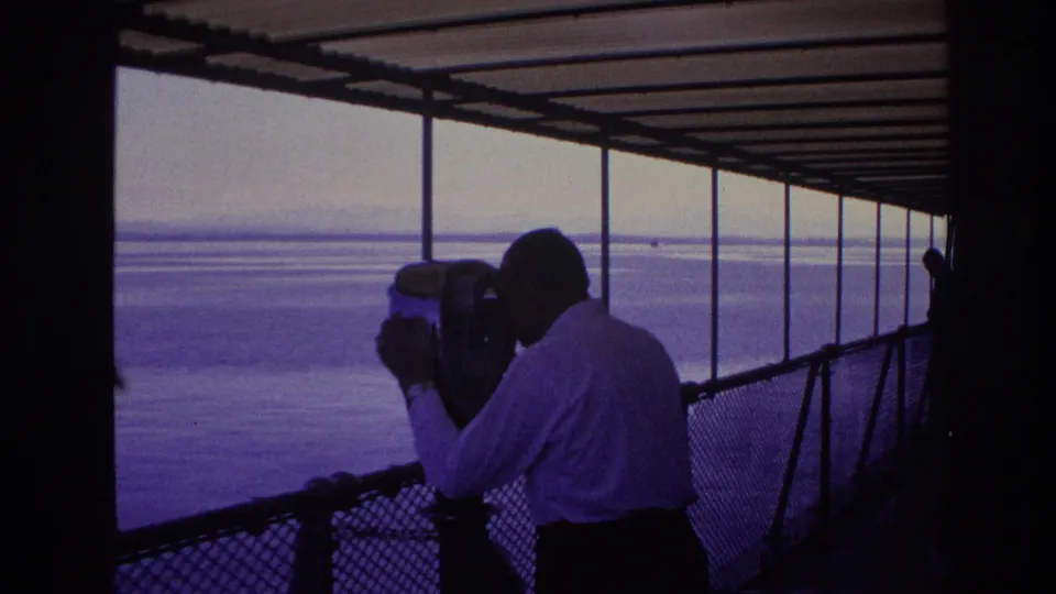 An adult man in silhouette peers through a public telescope from a covered waterfront structure, with open water stretching behind him.