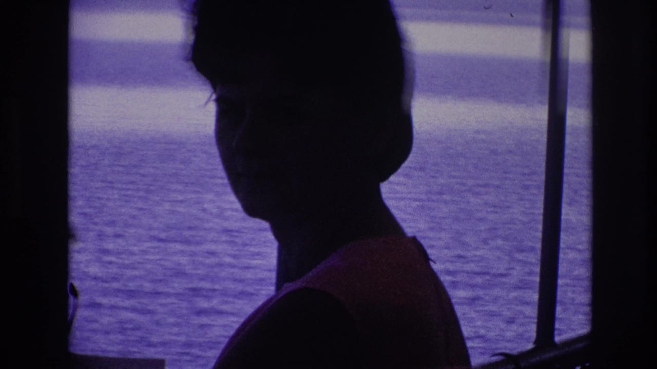 1967 8mm frame of a silhouetted woman by boat or dock railings, with open water and a distant shoreline in a purple color cast.