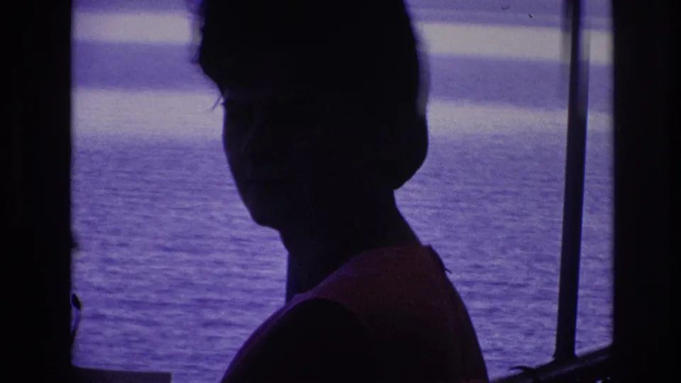 1967 8mm frame of a silhouetted woman by boat or dock railings, with open water and a distant shoreline in a purple color cast.