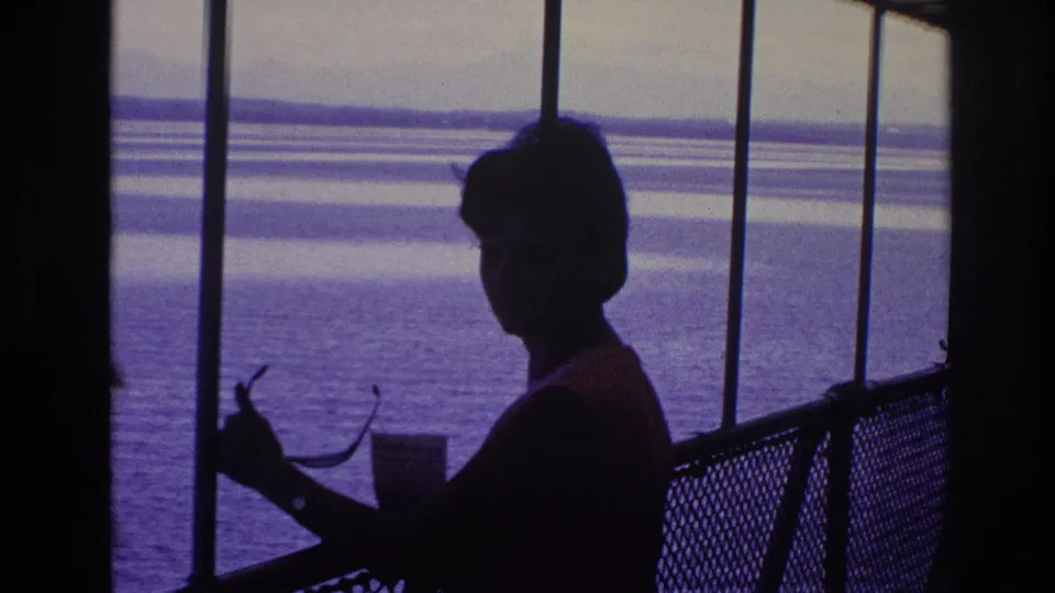 Home-movie frame showing a silhouetted woman by a mesh railing, holding sunglasses as open water and horizontal shoreline bands fill the background.