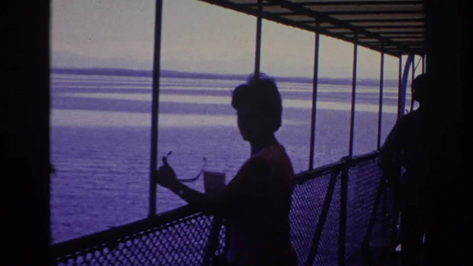 Wider 8mm view of a woman in silhouette under a canopy or deck structure, standing near railings with water and shoreline beyond.
