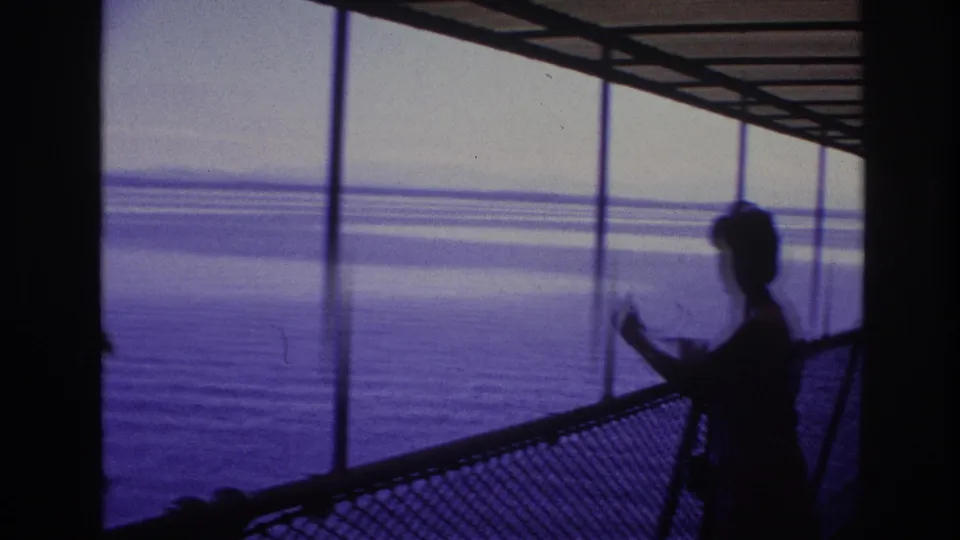 Silhouetted woman on a 1967 tour ship deck, framed by railings and vertical supports with water beyond.