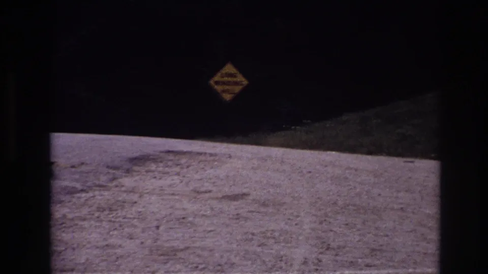 Dark 1967 travel frame with a yellow winding-road warning sign overlooking a pale gravel road and roadside slope.