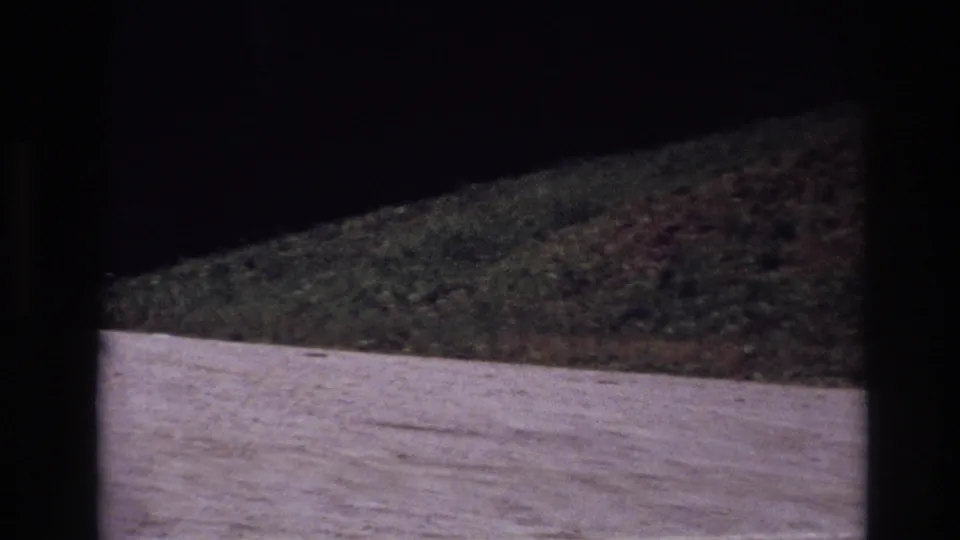 1967 Ottawa travel footage frame showing a pale gravel road curving beside a grassy embankment in low exposure.