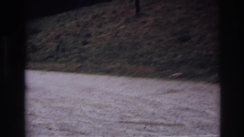 Dim 1967 Ottawa home-movie frame of a gravel road and grassy slope, with heavy shadow and visible film grain.