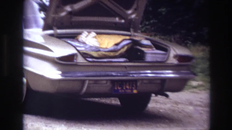 Handheld 8mm frame of a classic sedan's rear quarter, open trunk, packed blankets, and license plate along a dirt roadside in Ottawa.