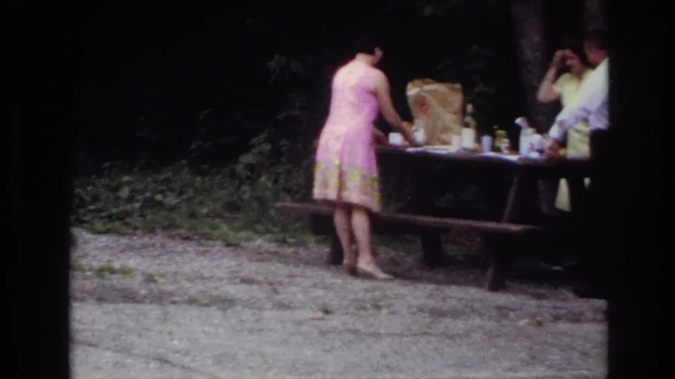 1967 Ottawa picnic scene with a woman in a pink dress arranging items while two adults stand nearby at a wooden table.