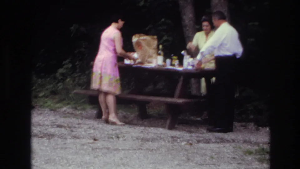 1967 Ottawa home-movie frame of adults at a wooded picnic table as a woman in a pink patterned dress handles items on the table.