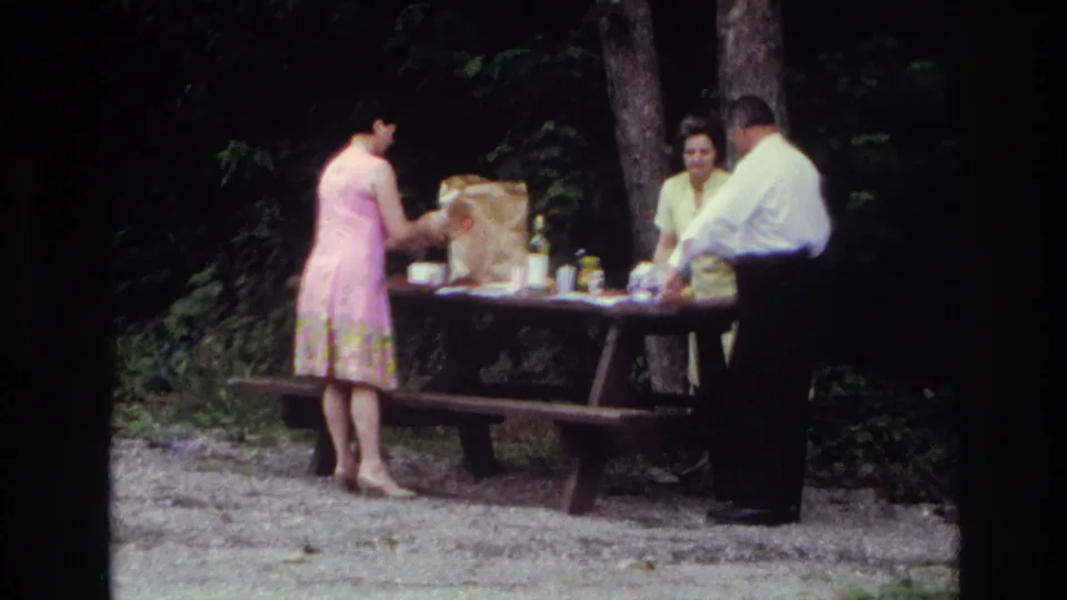 1967 Ottawa wooded picnic scene with three adults at a table holding food and drink items on a gravel surface.