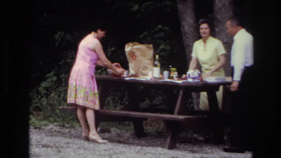 1967 Ottawa park picnic frame with a woman arranging food beside a brown paper bag while two adults stand near the table.