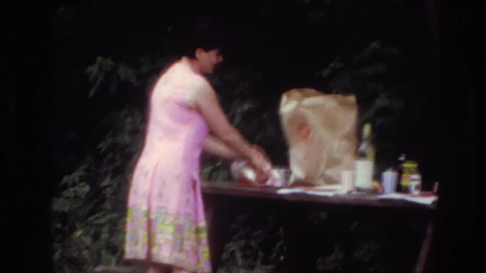 Closer 8mm frame from the 1967 Ottawa picnic, showing a woman in a pink patterned dress leaning over food and supplies.