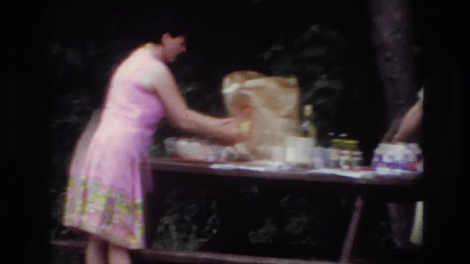 1967 Ottawa home-movie frame showing a woman reaching across a picnic table with bottles, cans and a brown paper bag visible.