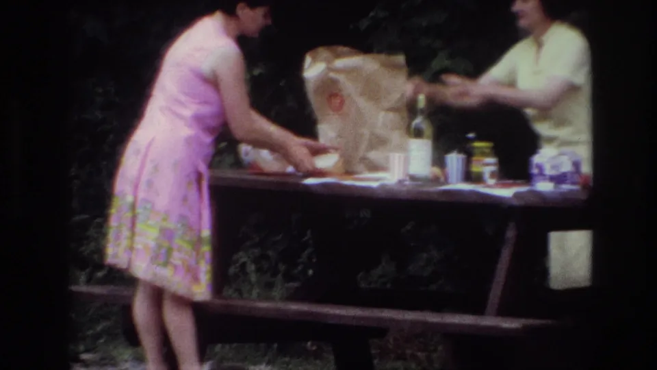 In the supplied 1967 Ottawa home-movie context, a woman in a pink dress arranges items on a wooded picnic table.