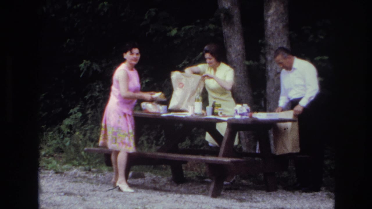 Two women and a man stand around a picnic table in a wooded area.