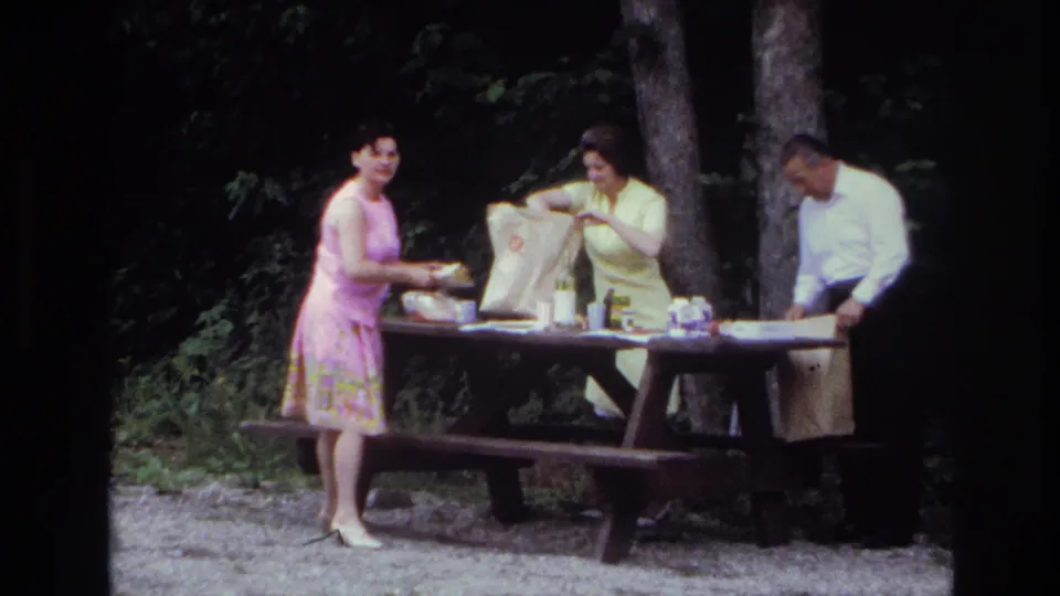 Three adults gather at a picnic table in the supplied 1967 Ottawa park setting, unpacking food from a large paper bag.