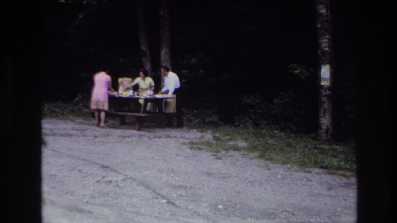 1967 Ottawa home-movie frame showing three adults preparing a picnic at a wooden table near dark trees.
