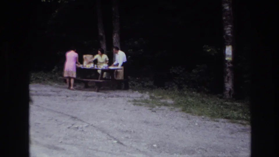 1967 Ottawa home-movie frame showing three adults preparing a picnic at a wooden table near dark trees.