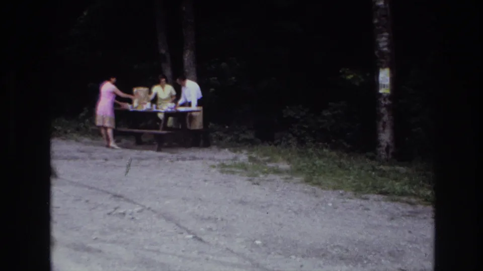 1967 Ottawa picnic scene with a woman in a pink dress reaching across a wooden table as two adults handle supplies.