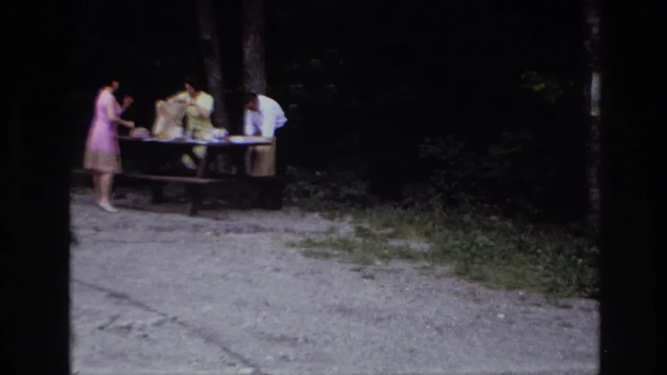 Home-movie frame from Ottawa in 1967 showing adults organizing picnic items on a table in a wooded clearing.