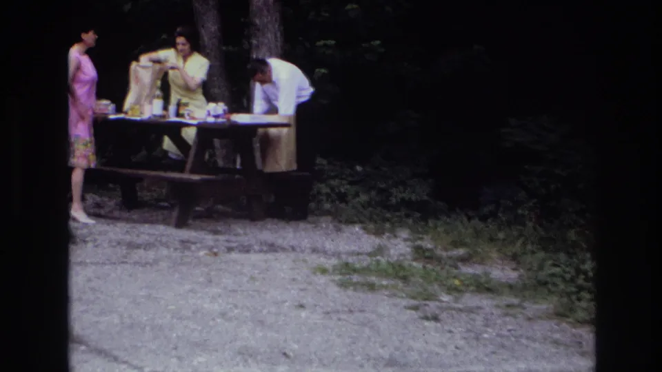 1967 Ottawa 8mm frame showing a closer view of adults unpacking picnic supplies at a wooden table near trees.