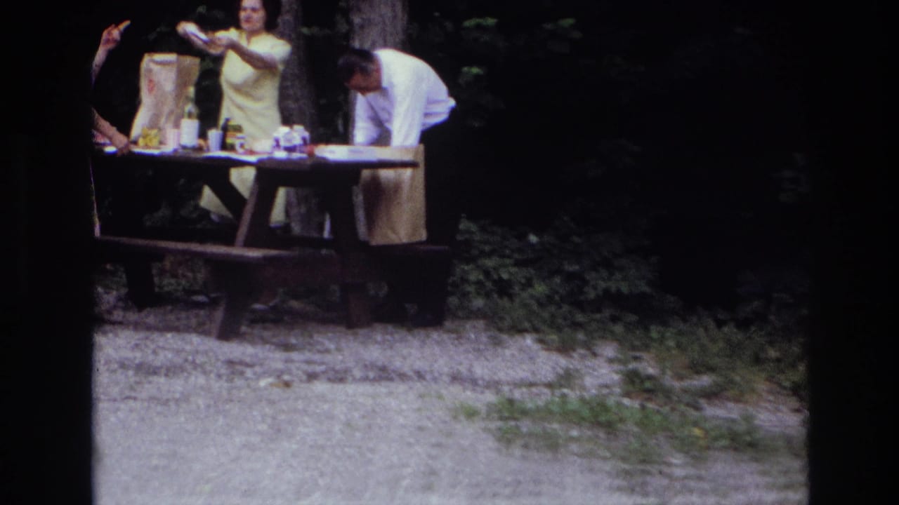 1967 Ottawa home-movie frame showing adults unpacking picnic items at a wooden table in a shaded park.