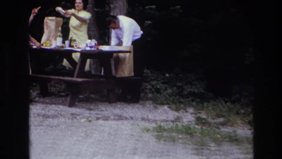 1967 Ottawa home-movie frame showing adults unpacking picnic items at a wooden table in a shaded park.