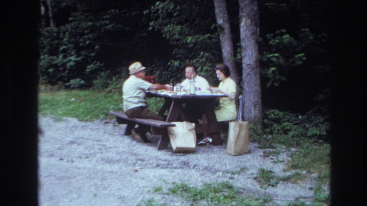 Three adults sit at a wooden picnic table in a gravel clearing beside trees.