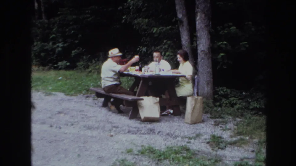 At the wooded Ottawa picnic table, one man raises a drink while two other adults sit nearby with bottles and bags on the ground.