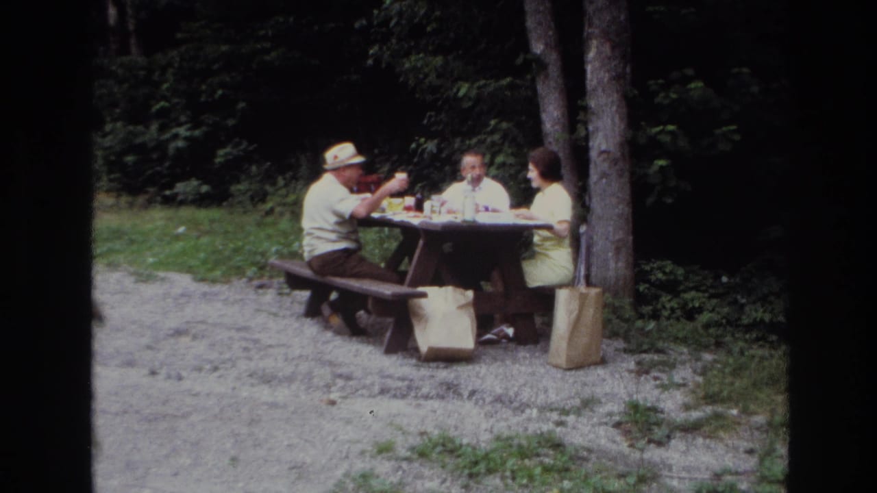 1967 Ottawa home-movie frame showing three adults at a wooden picnic table beside trees and brown paper bags.