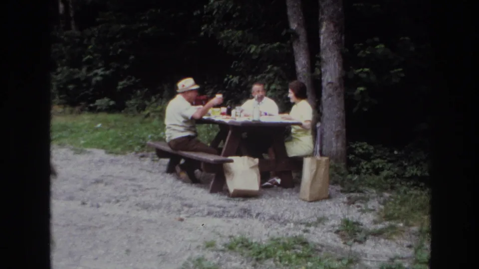 1967 Ottawa home-movie frame showing three adults at a wooden picnic table beside trees and brown paper bags.