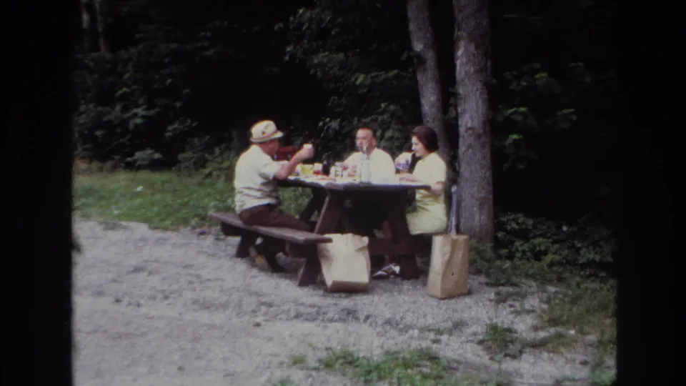 Home-movie frame from Ottawa in 1967 showing a family picnic conversation at a wooden table in the woods.