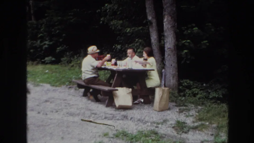 1967 Ottawa 8mm frame of three adults eating or talking at a wooded picnic table with bags on the ground.