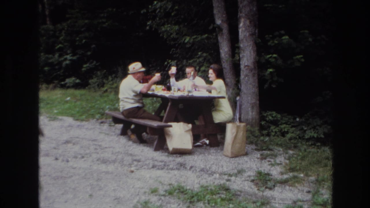 In 1967 Ottawa home-movie footage, three adults sit at a picnic table with food and drinks near trees and gravel ground.