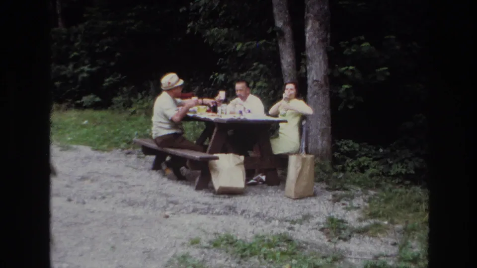 Three adults continue a park picnic in Ottawa, with glasses and lunch items on the table and paper bags beside the benches.
