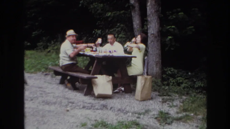 The home-movie frame shows a man in a light hat at left facing two other adults during a wooded Ottawa picnic lunch.