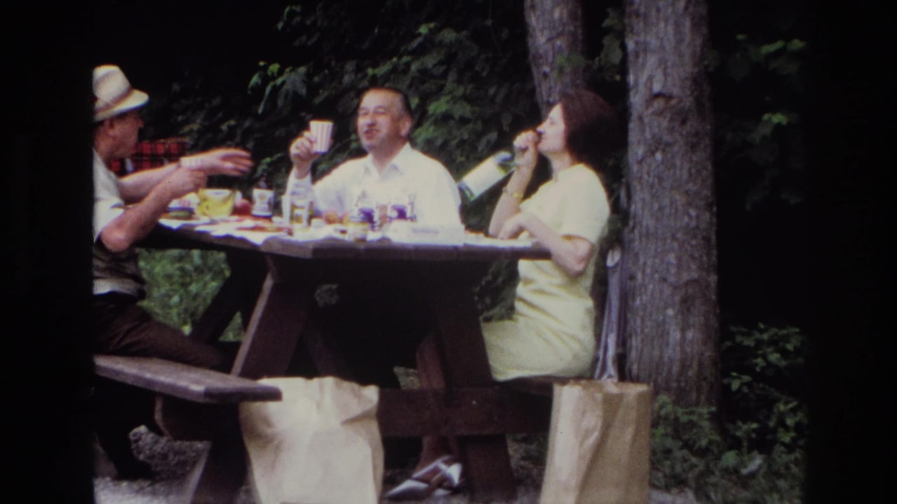 1967 Ottawa home-movie frame showing adults at a wooded picnic table with cups, bottles, and a woman in a yellow dress.