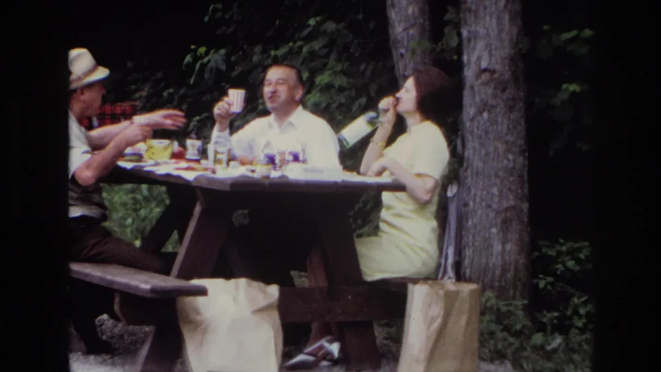 1967 Ottawa home-movie frame showing adults at a wooded picnic table with cups, bottles, and a woman in a yellow dress.