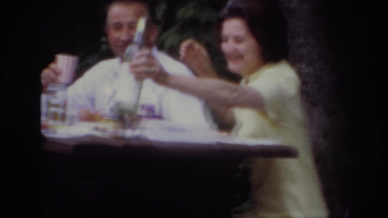 1967 Ottawa home-movie frame of a woman in yellow smiling as she raises a bottle toward the camera at a wooded picnic table.
