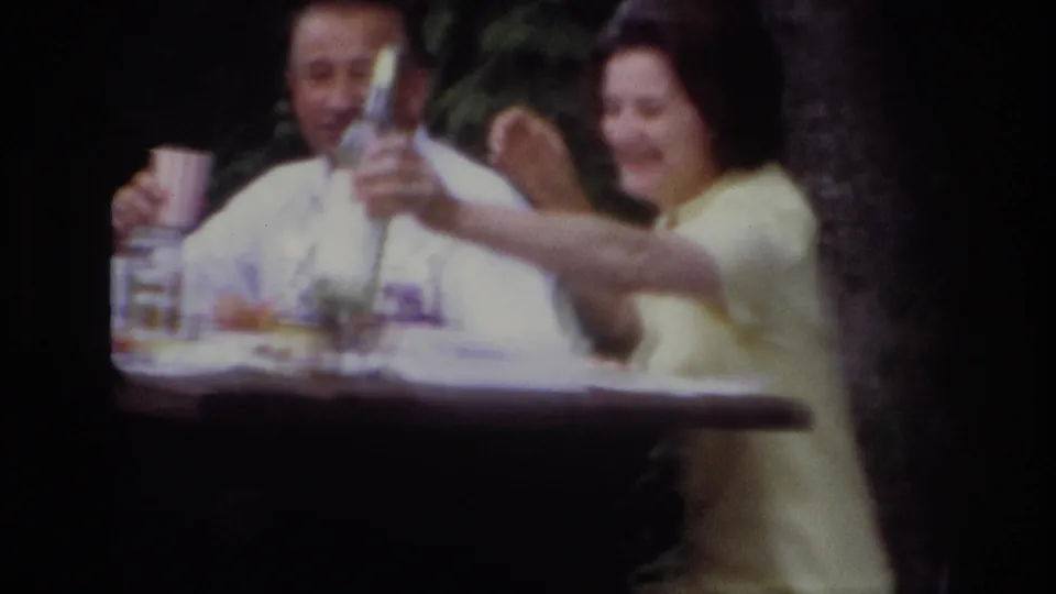 1967 Ottawa home-movie frame of a woman in yellow smiling as she raises a bottle toward the camera at a wooded picnic table.