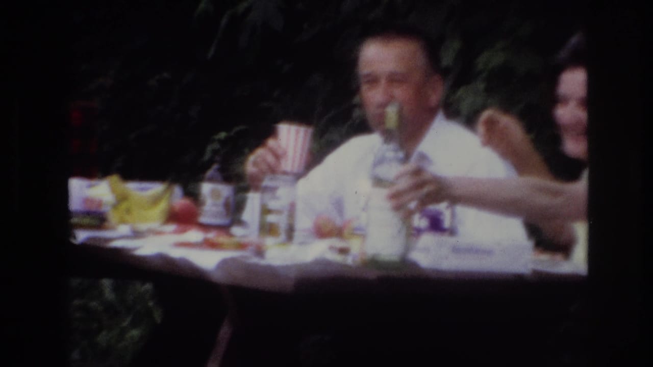 1967 Ottawa home-movie frame of adults around a picnic table, with a man in a light shirt holding a cup near a bottle and fruit.