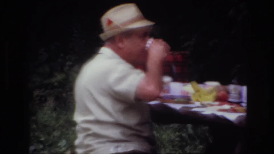 Side view from 1967 Ottawa home-movie footage showing a man in a straw hat drinking at a picnic table with fruit and tableware.