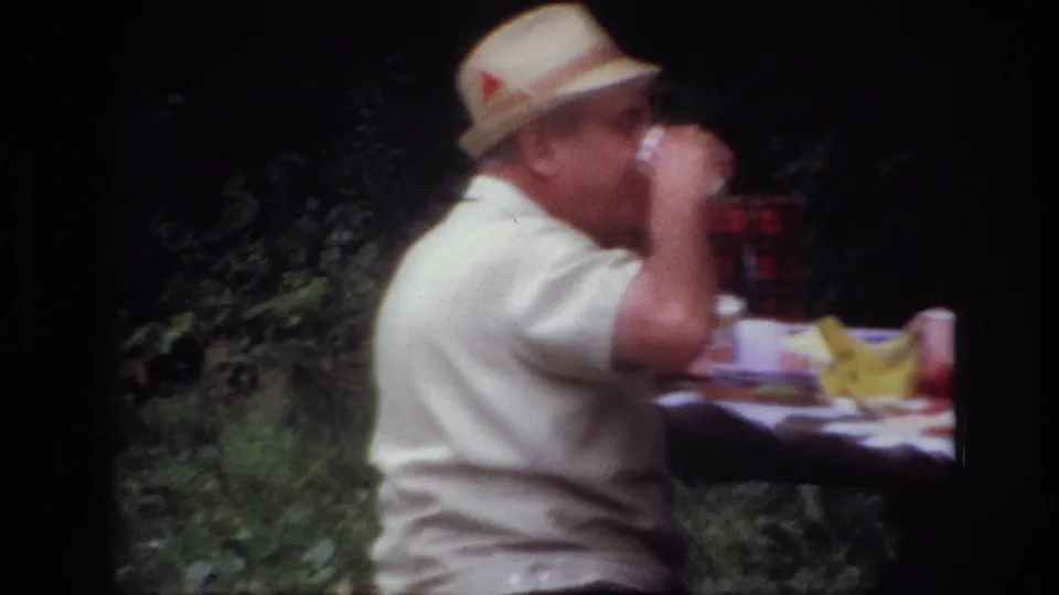 1967 Ottawa frame of a seated man in a straw hat drinking from a cup, with picnic food and foliage in the background.