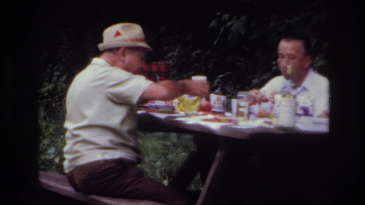 Two adult men sit across a picnic table in wooded Ottawa home-movie footage, with cups, food, and a glass bottle visible.