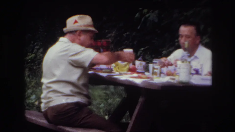 Two adult men sit across a picnic table in wooded Ottawa home-movie footage, with cups, food, and a glass bottle visible.