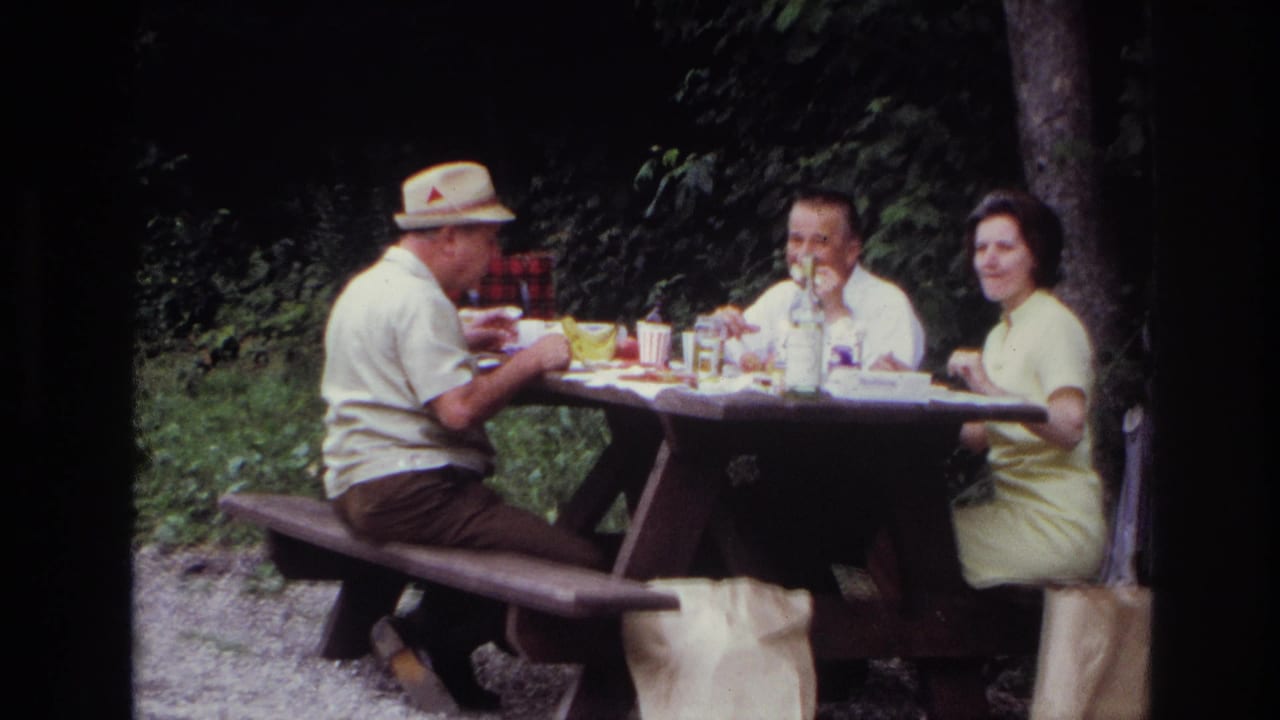 Three adults sit at a wooden picnic table in Ottawa in 1967, with food, cups, a bottle, and trees around them.