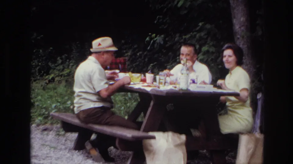 Three adults sit at a wooden picnic table in Ottawa in 1967, with food, cups, a bottle, and trees around them.