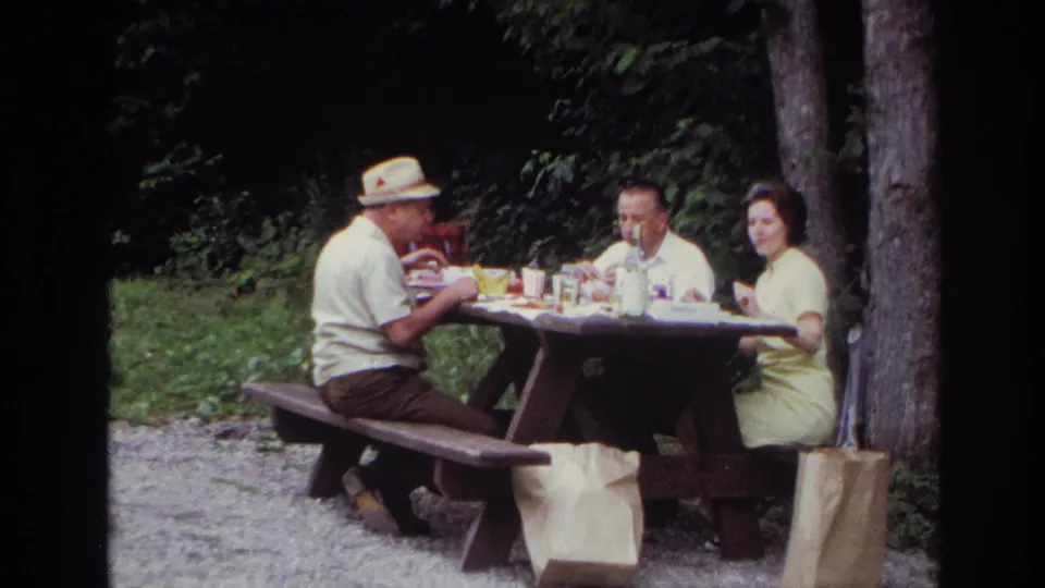 Ottawa 1967 home-movie frame showing three adults eating at a wooden picnic table in a forested park setting.